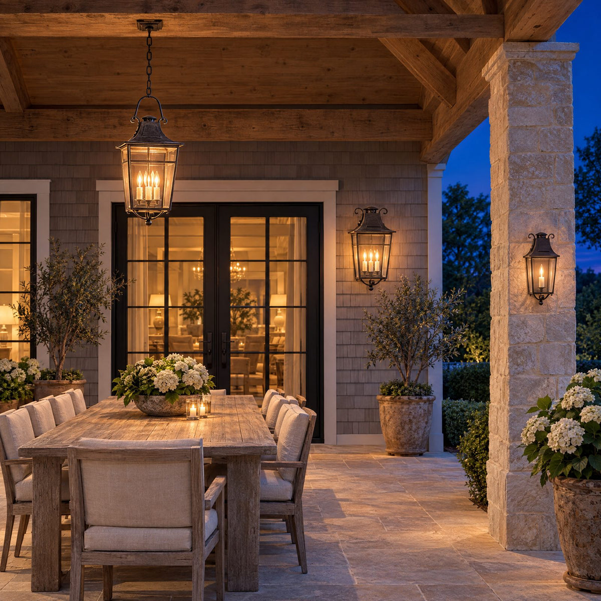 Shop the look: Covered limestone patio at dusk with a rust-iron hanging lantern over a teak dining table, matching wall sconces flanking French doors, and a post light at the garden steps
