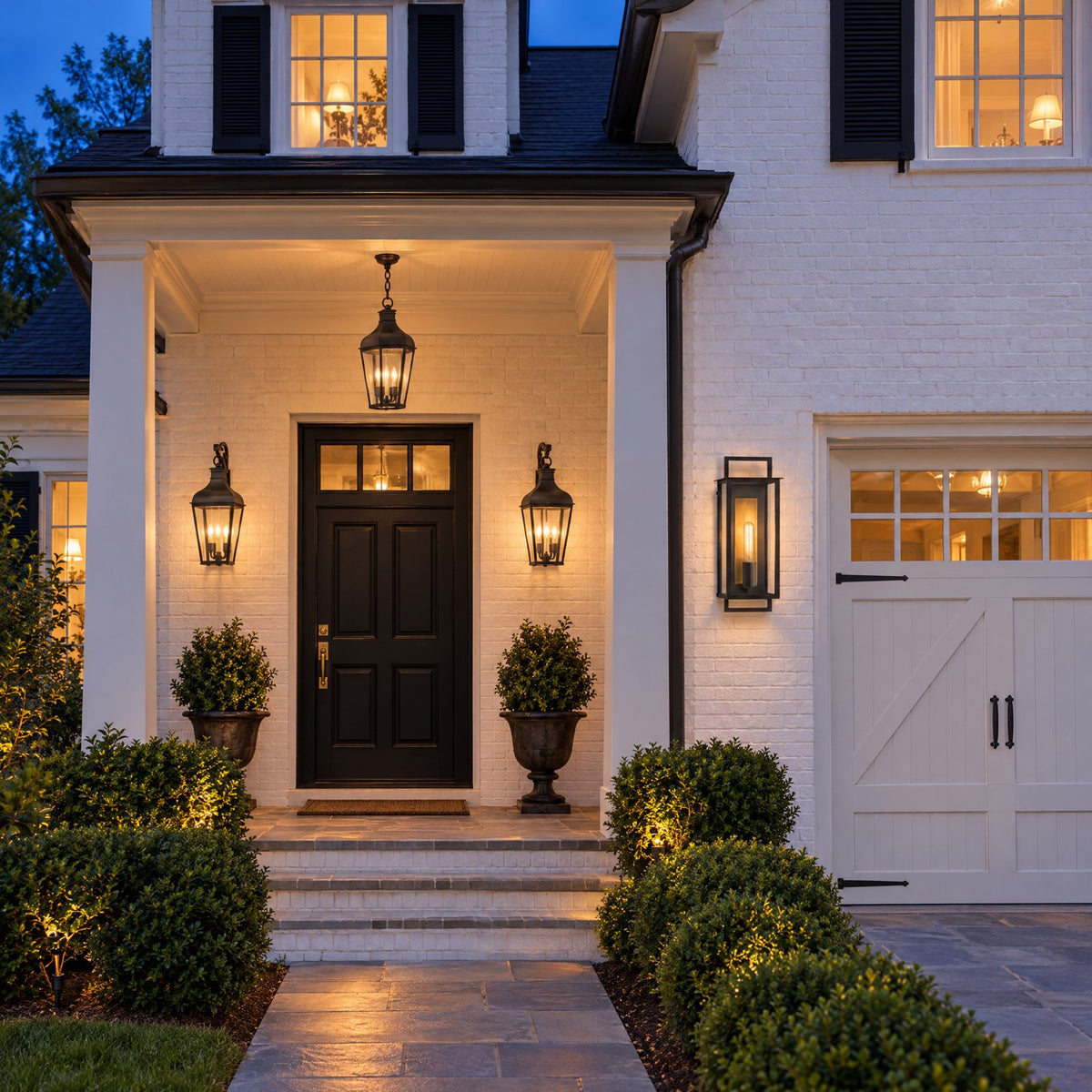 Shop the look: White brick house at dusk with rust-iron bracketed lanterns flanking the front door, a matching hanging porch lantern, and aged-iron wall lanterns at the garage